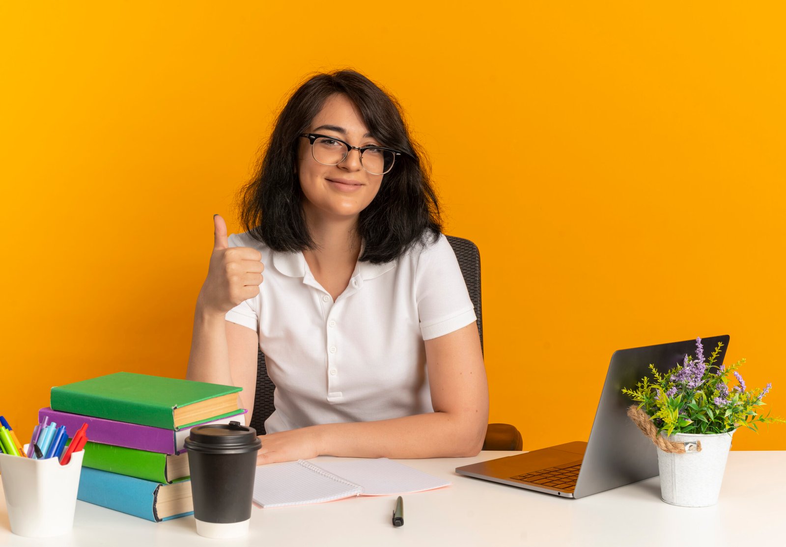 young pleased pretty caucasian schoolgirl wearing glasses sits at desk with school tools thumbs up isolated on orange background with copy space